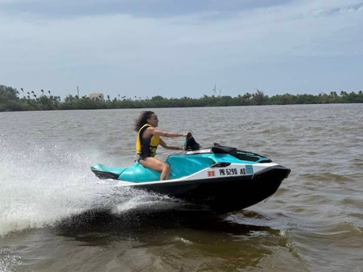 Person riding a teal jet ski on a lake with trees in the background.