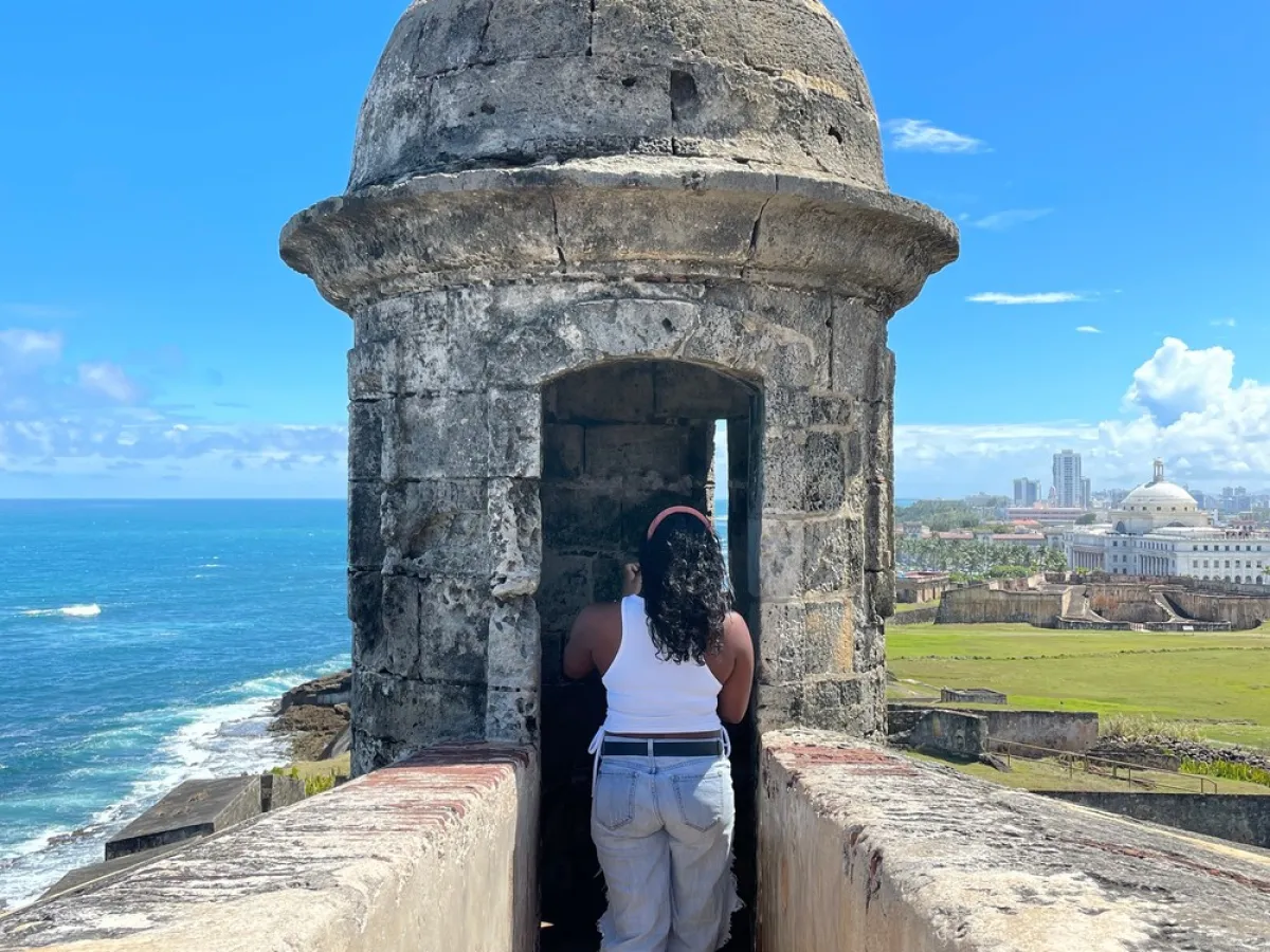 Person standing in stone fort turret overlooking ocean and cityscape under clear blue sky.