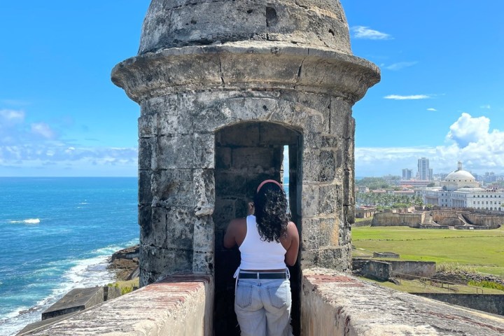 Person standing in stone fort turret overlooking ocean and cityscape under clear blue sky.