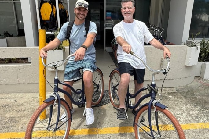 Two men pose with bikes outside a building, smiling at the camera.