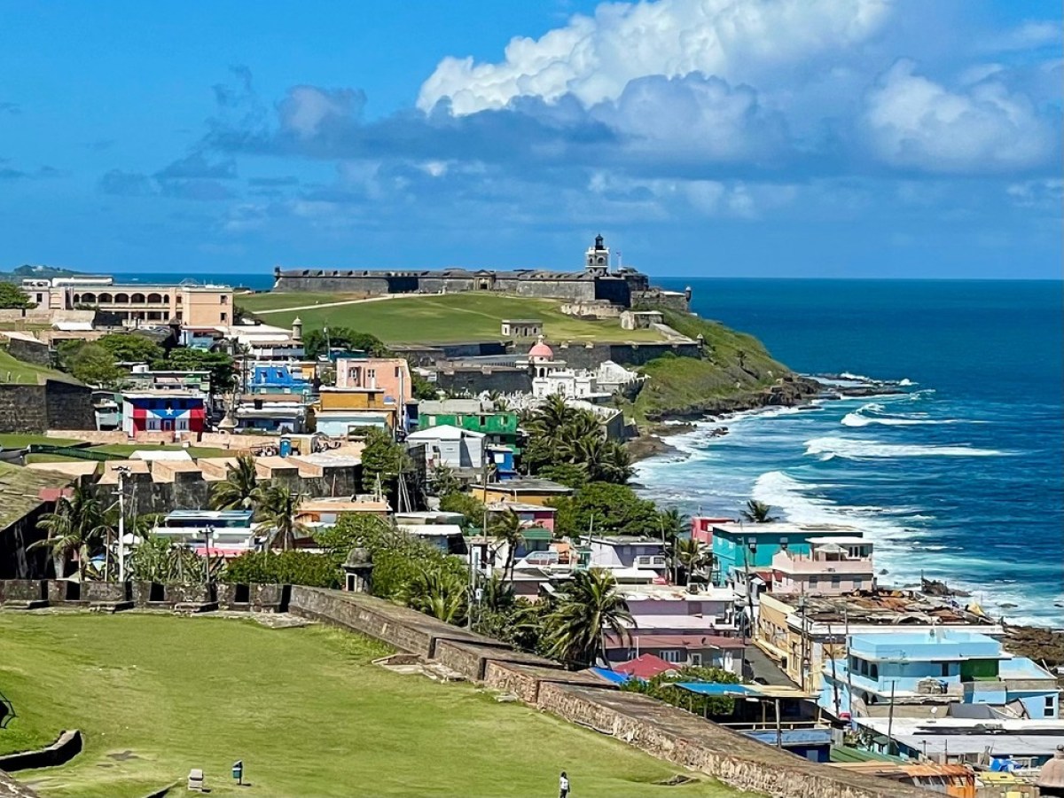 Colorful coastal town with fort, palm trees, and waves under a blue sky with clouds.