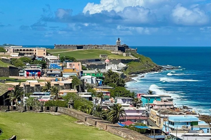 Colorful coastal town with fort, palm trees, and waves under a blue sky with clouds.