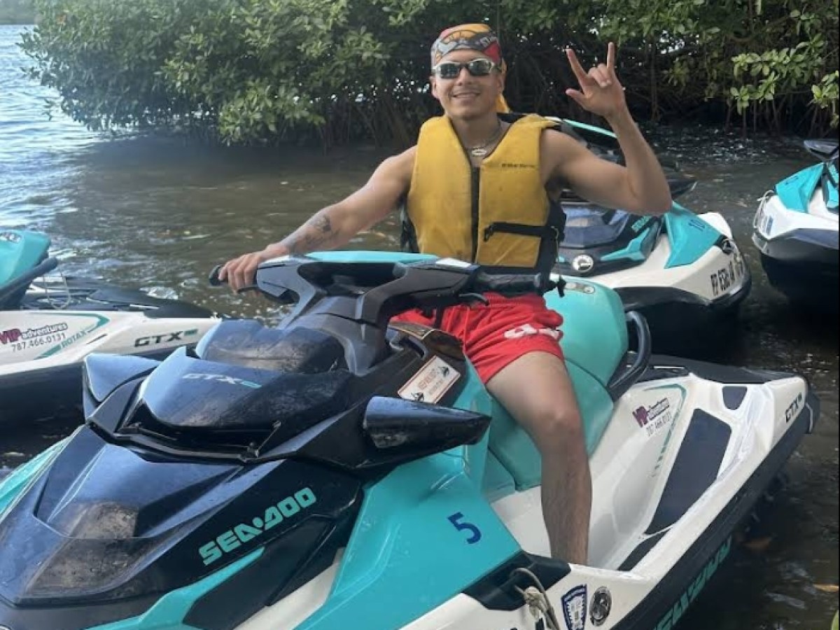 Person on a jet ski wearing a yellow life vest, making a hand sign near water and trees.