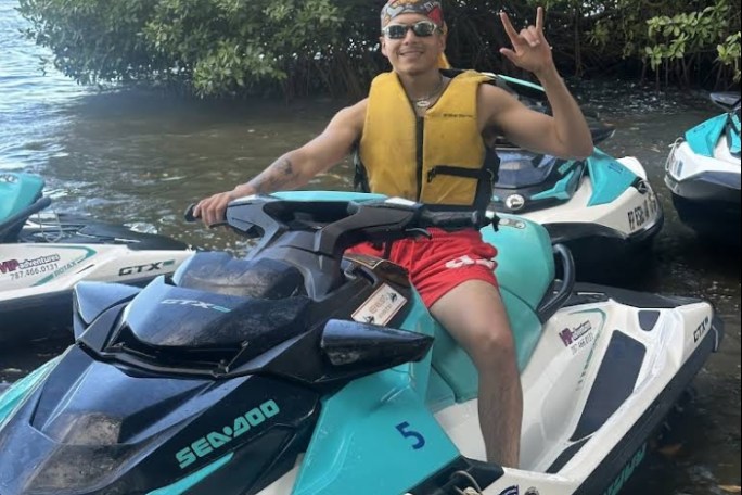Person on a jet ski wearing a yellow life vest, making a hand sign near water and trees.