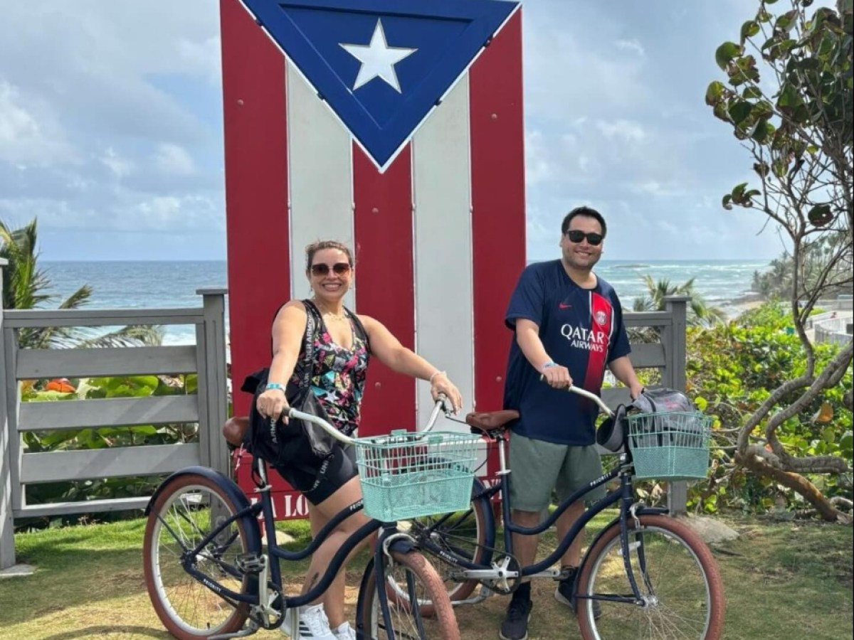 Two people with bikes in front of a large Puerto Rican flag near the ocean.
