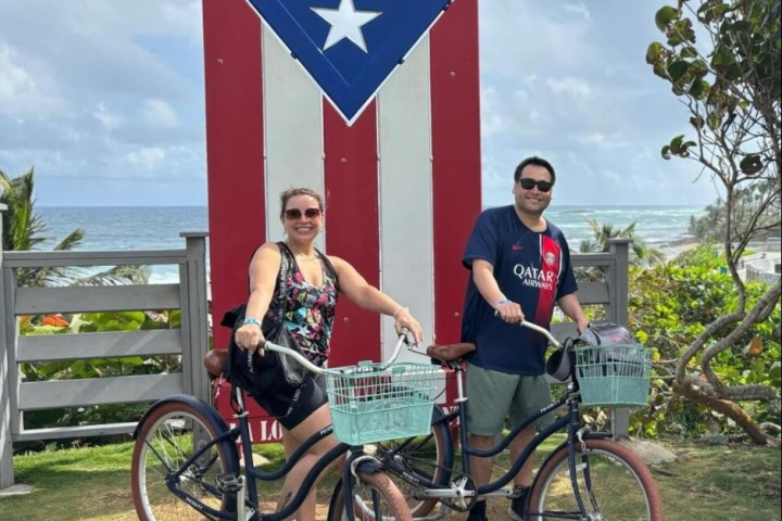 Two people with bikes in front of a large Puerto Rican flag near the ocean.