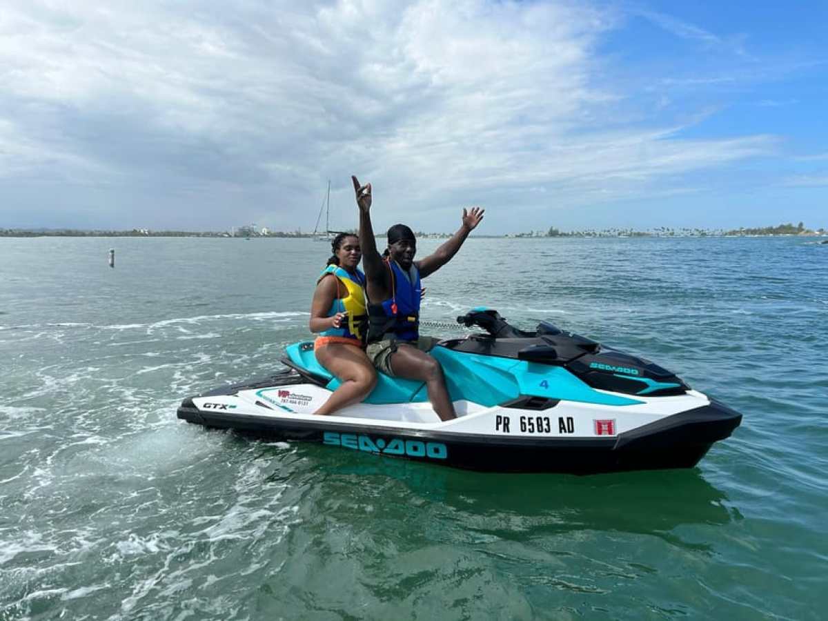 Two people on a jet ski wearing life vests, waving on a sunny day.