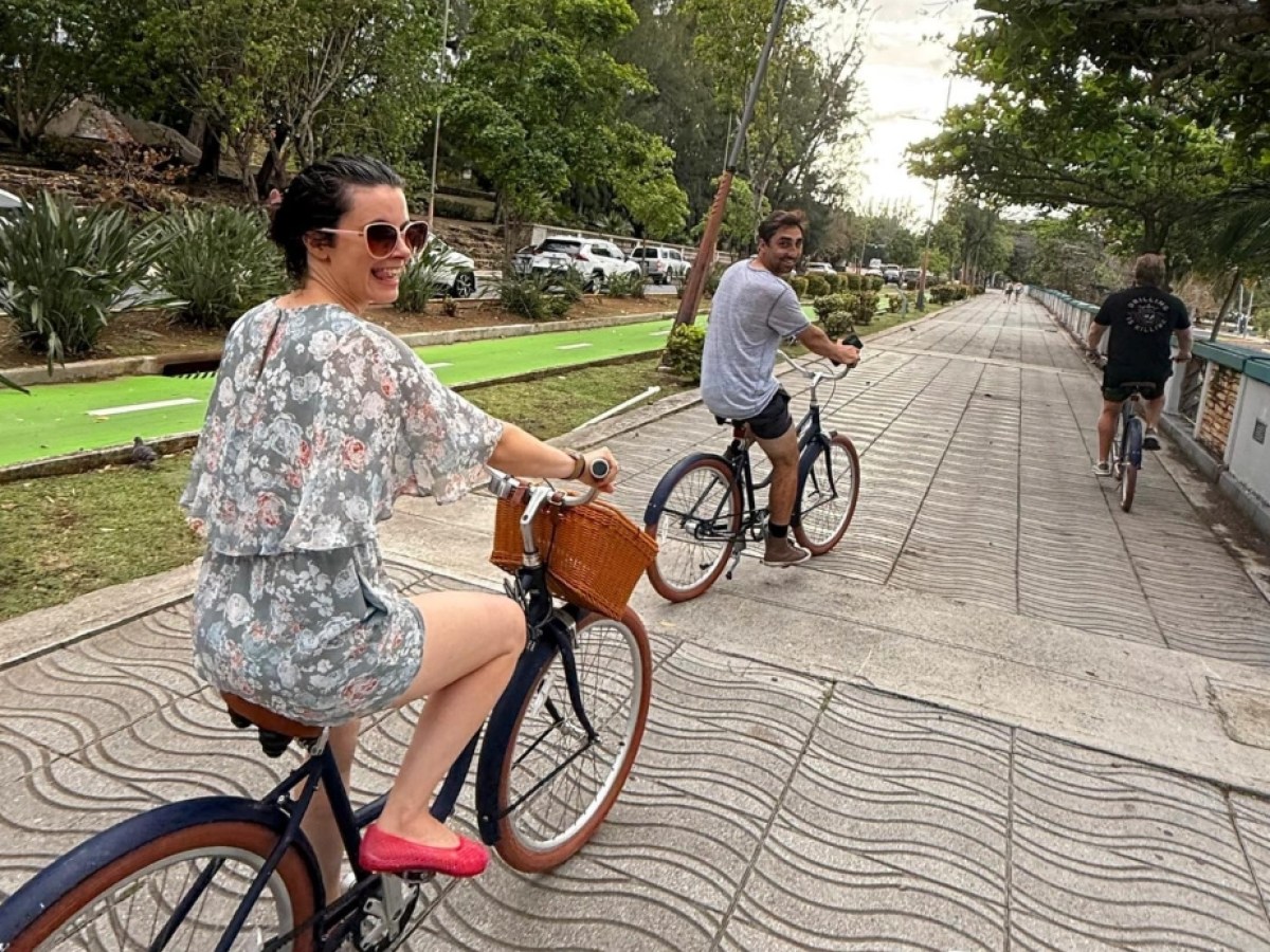 Three people biking on a tree-lined path, two facing back, one wearing floral dress and red shoes.