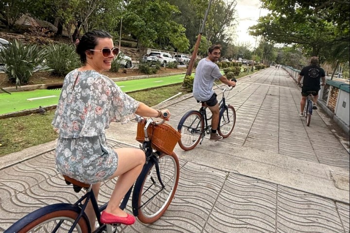 Three people biking on a tree-lined path, two facing back, one wearing floral dress and red shoes.
