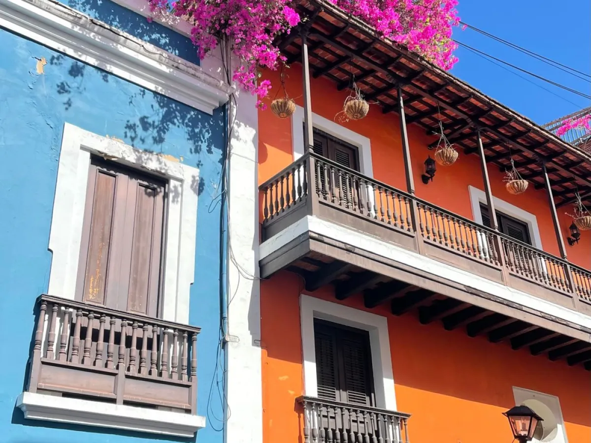 Colorful blue and orange buildings with wooden balconies under pink flowers and a clear blue sky.