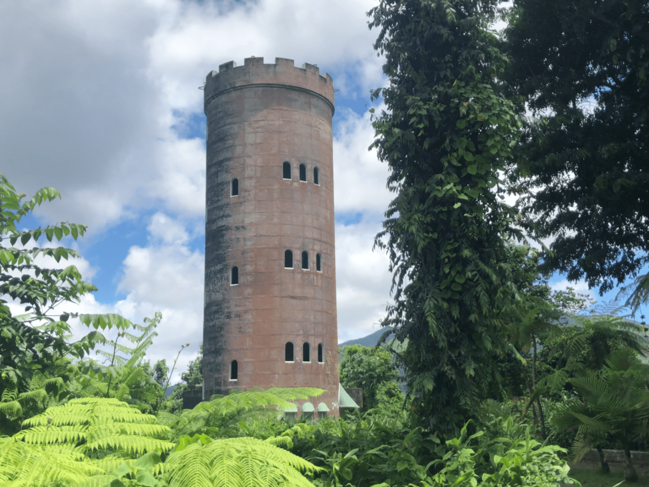 Tall stone tower with small windows, surrounded by lush green foliage under a partly cloudy sky.