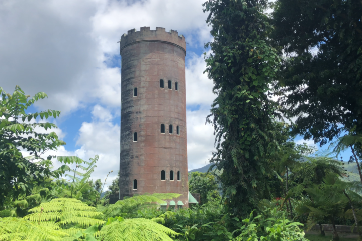 Tall stone tower with small windows, surrounded by lush green foliage under a partly cloudy sky.
