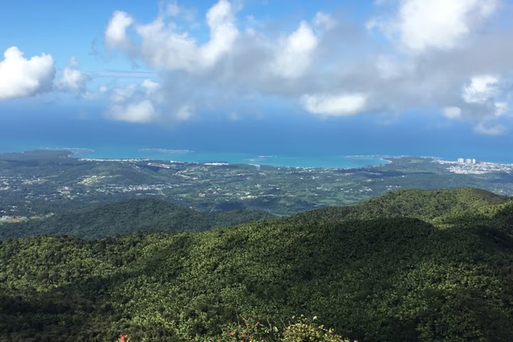 Aerial view of lush green hills with a coastline in the distance under a partly cloudy blue sky.