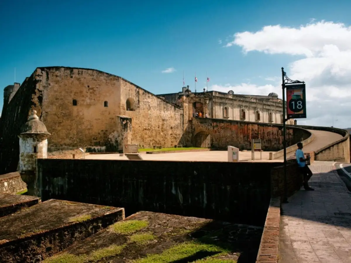 Stone fortress with curved walkway under a blue sky, person walking, sign with number 18.