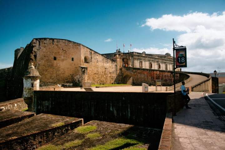 Stone fortress with curved walkway under a blue sky, person walking, sign with number 18.