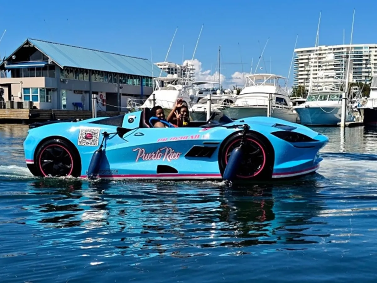 A turquoise car-shaped boat on water near a marina with yachts and buildings in the background.