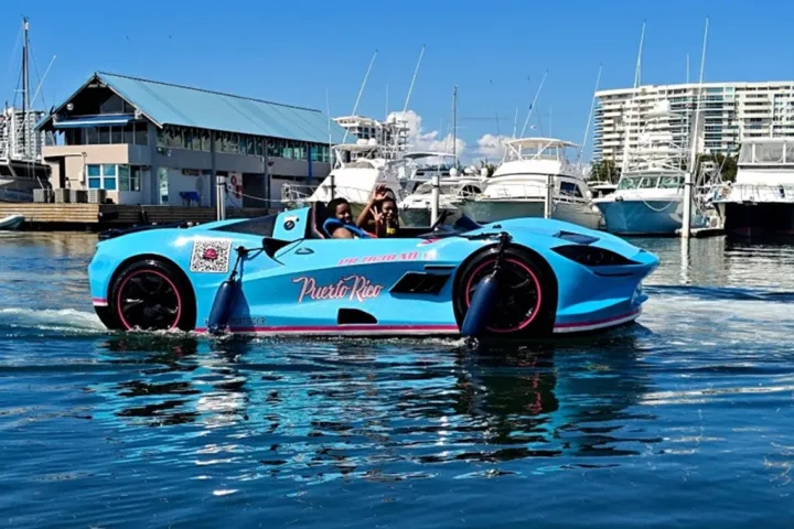 A turquoise car-shaped boat on water near a marina with yachts and buildings in the background.