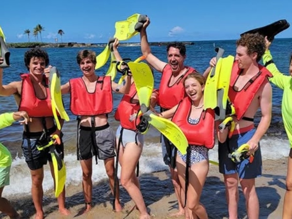Group of people on a beach wearing red life vests, holding snorkeling gear, smiling and posing together.