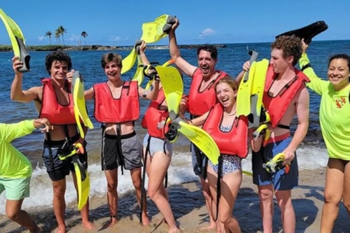 Group of people on a beach wearing red life vests, holding snorkeling gear, smiling and posing together.