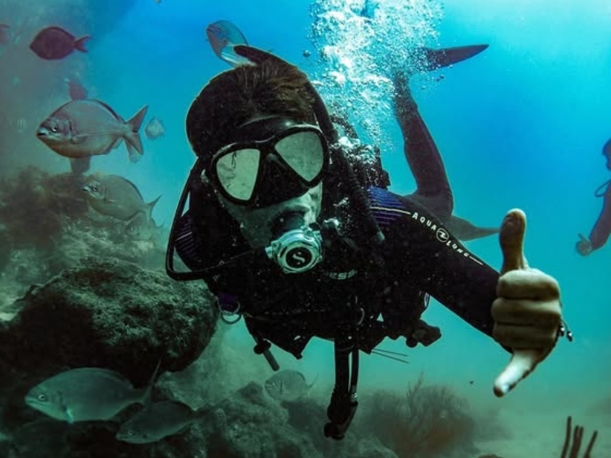 Scuba diver underwater giving thumbs up surrounded by fish.