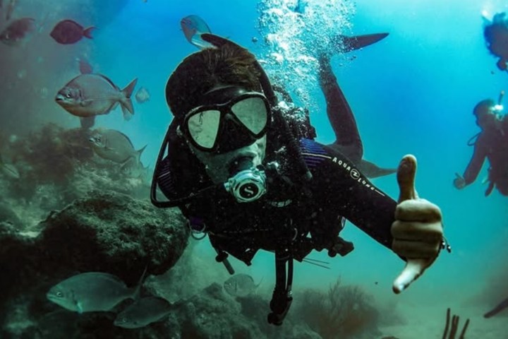 Scuba diver underwater giving thumbs up surrounded by fish.