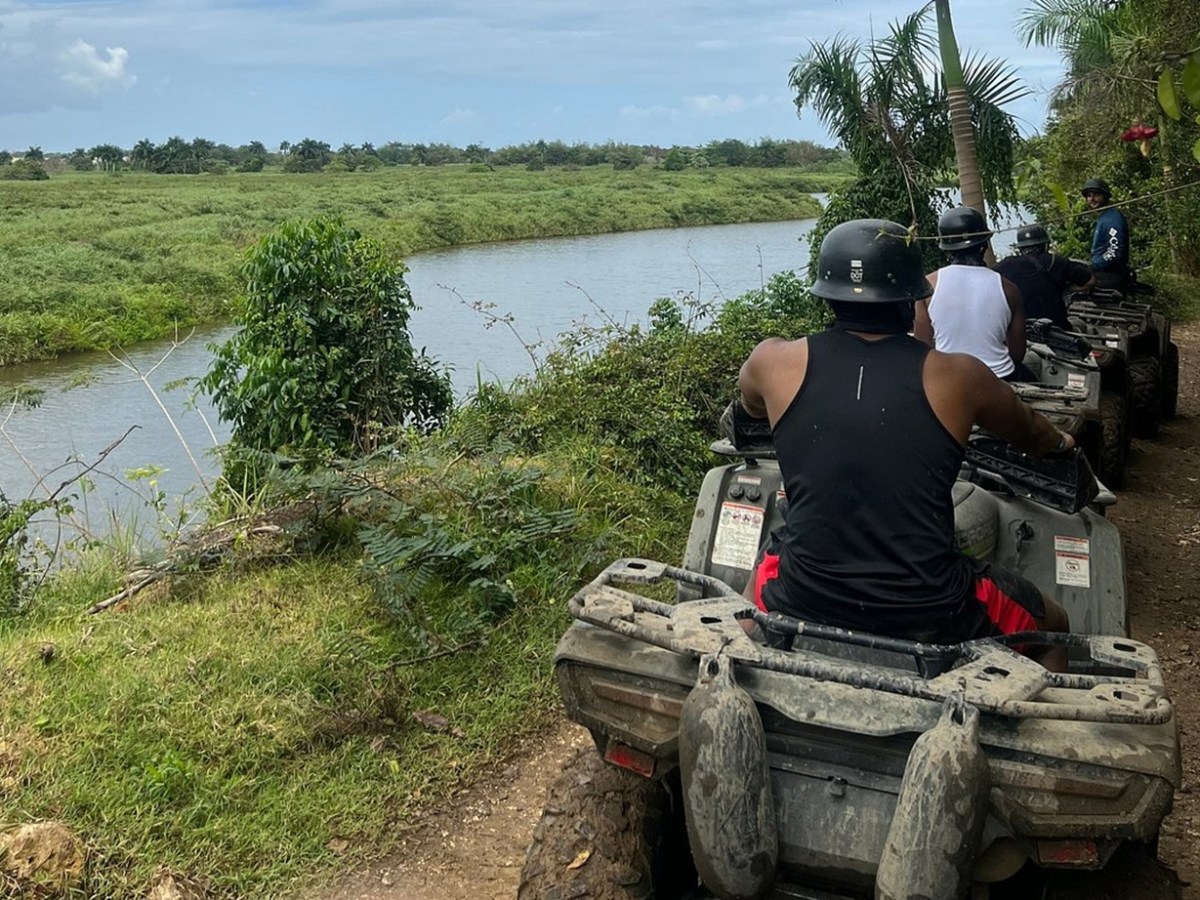 Group riding ATVs along a riverbank trail with lush greenery and palm trees.