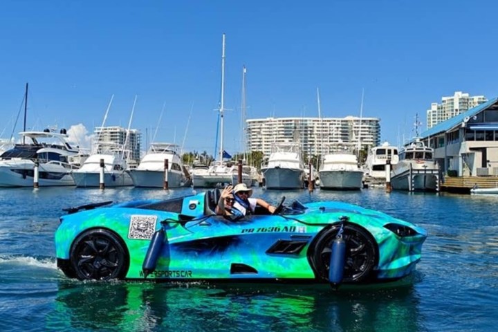 Blue amphibious car with two people driving on water near marina with boats and buildings.