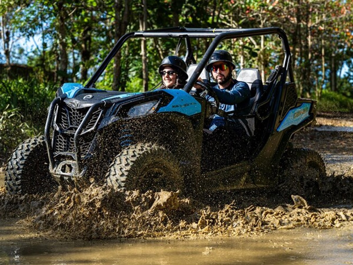 Two people in helmets driving a blue ATV through a muddy trail with trees in the background.