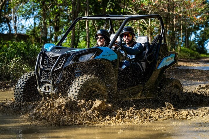 Two people in helmets driving a blue ATV through a muddy trail with trees in the background.