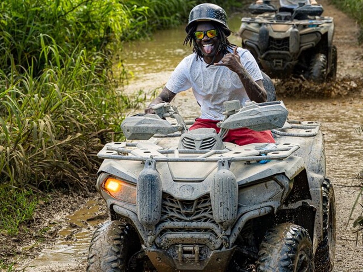 Person riding an ATV through a muddy path, wearing a helmet and sunglasses, with vegetation on both sides.