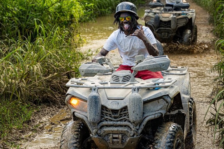 Person riding an ATV through a muddy path, wearing a helmet and sunglasses, with vegetation on both sides.