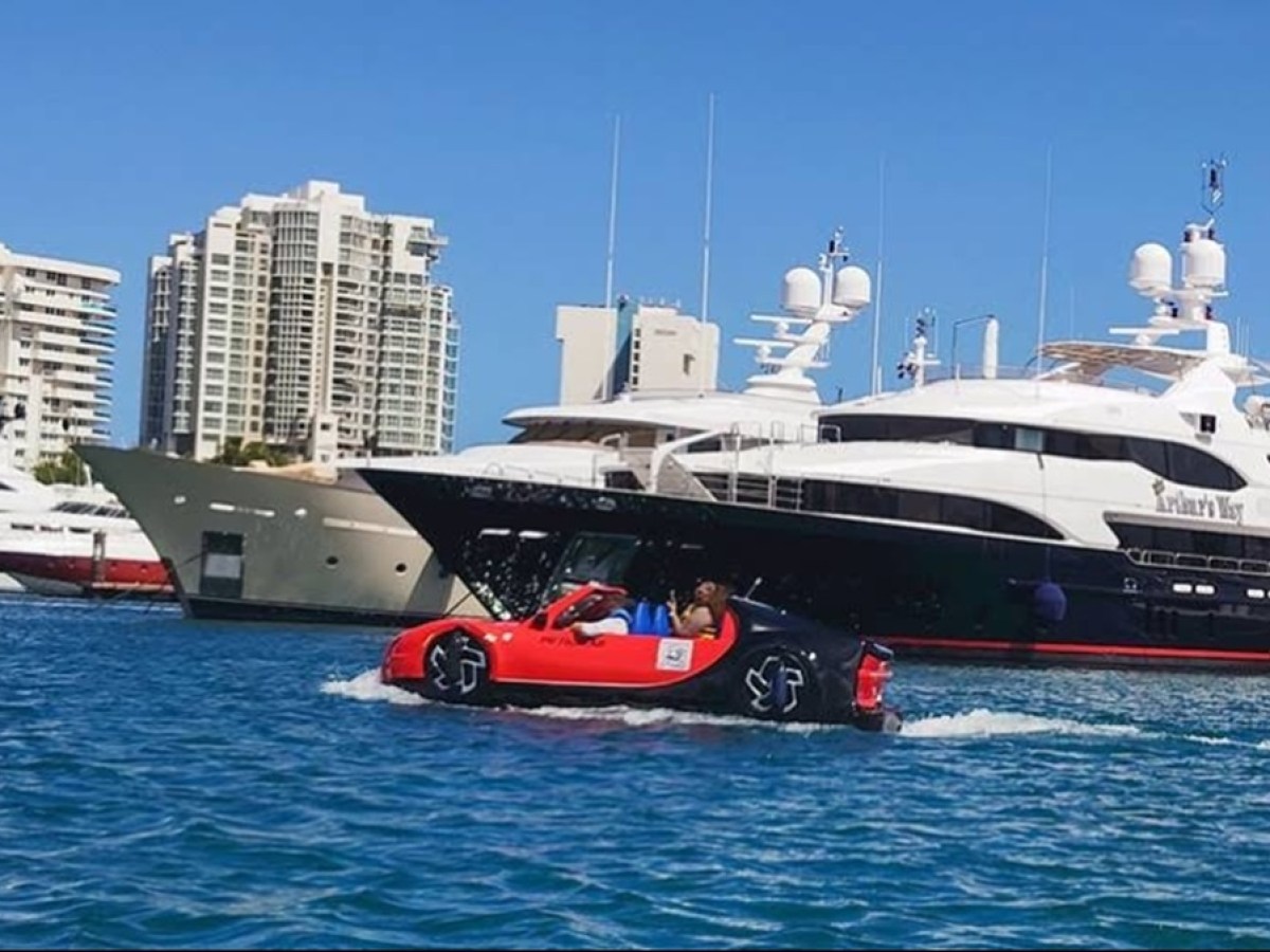 Red amphibious car on water near luxury yachts and tall buildings on a sunny day.