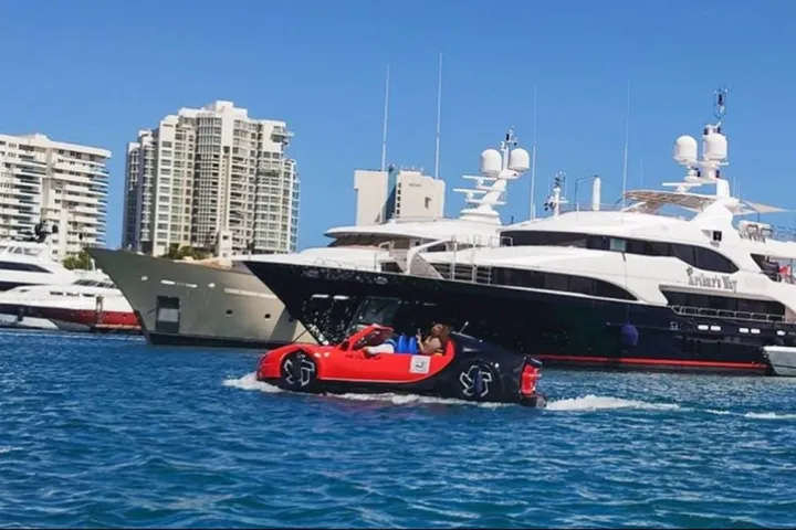 Red amphibious car on water near luxury yachts and tall buildings on a sunny day.