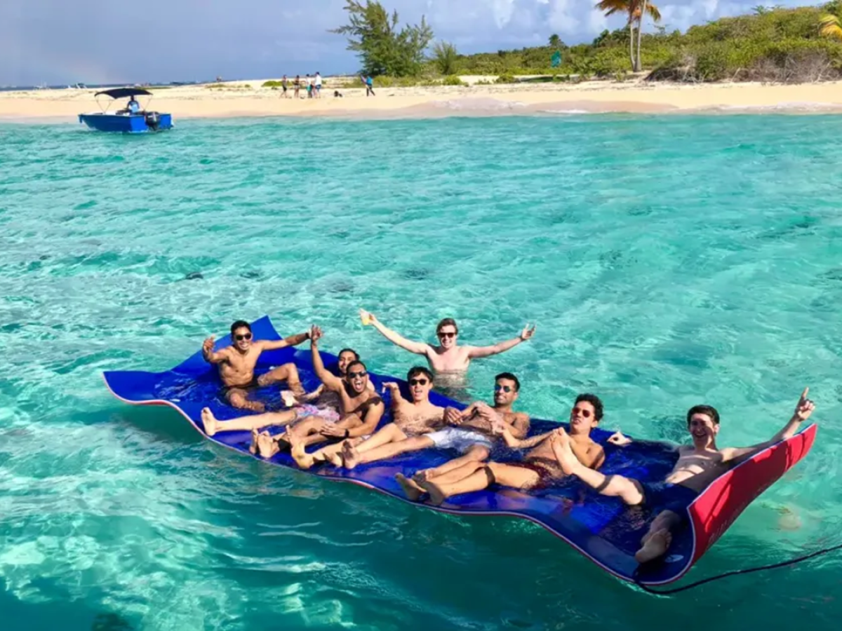 Group of people relaxing on a floating mat in clear turquoise water near a tropical beach.