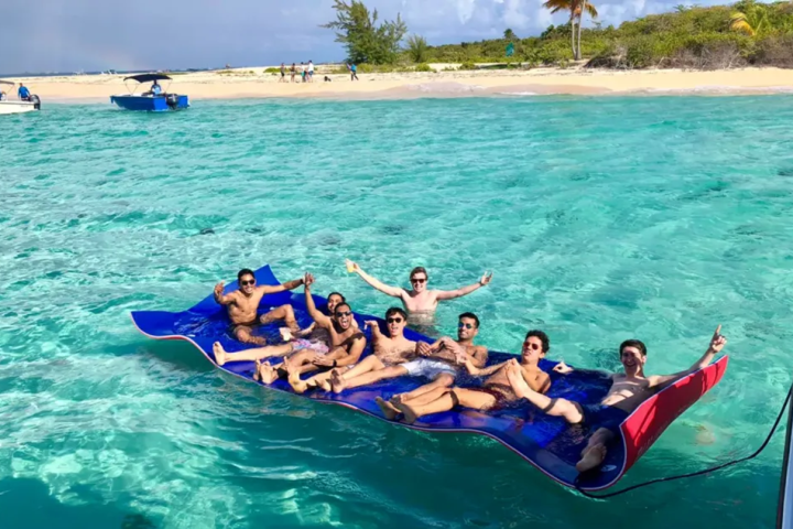 Group of people relaxing on a floating mat in clear turquoise water near a tropical beach.