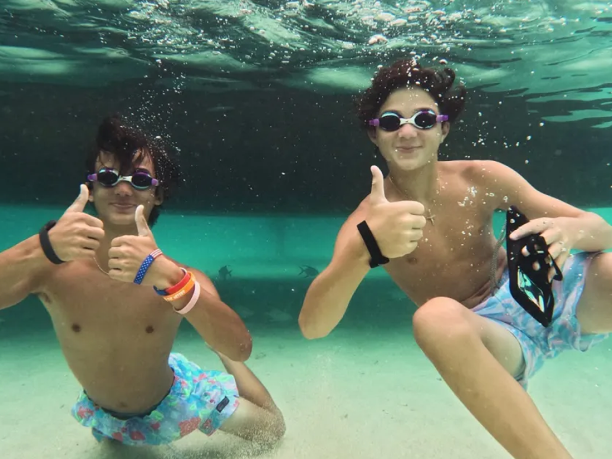 Two people underwater giving thumbs up, wearing goggles and swim trunks.