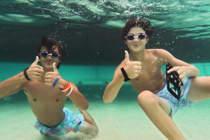 Two people underwater giving thumbs up, wearing goggles and swim trunks.