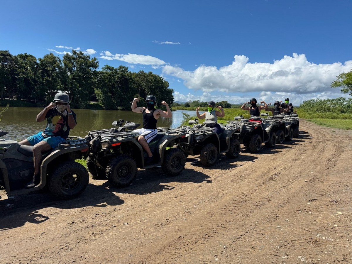 Group on ATVs flexing by a river under a blue sky with clouds and trees in the background.