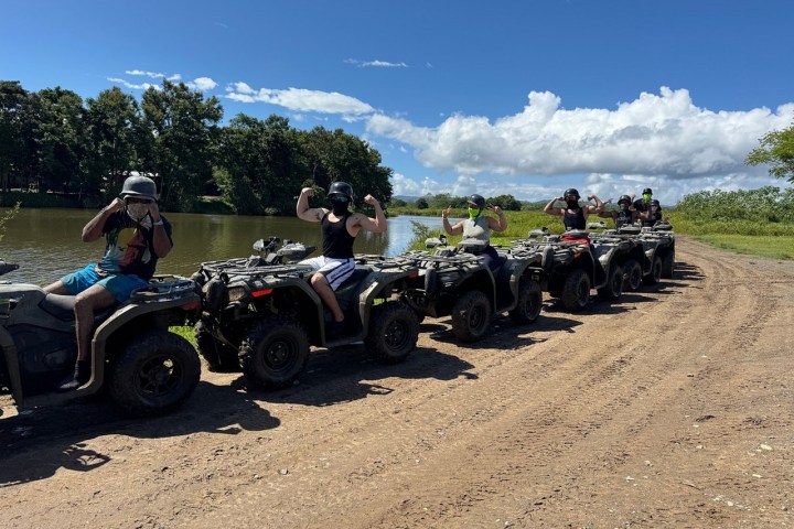Group on ATVs flexing by a river under a blue sky with clouds and trees in the background.