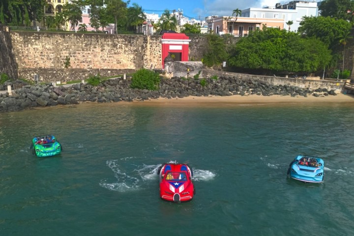 Three colorful boat cars on water near a historic stone fort and small beach.