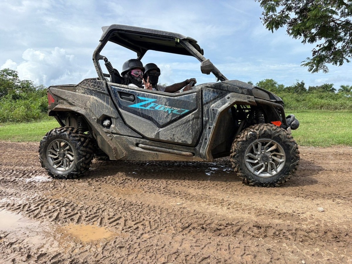 Two people in helmets driving a muddy off-road vehicle on a dirt path.