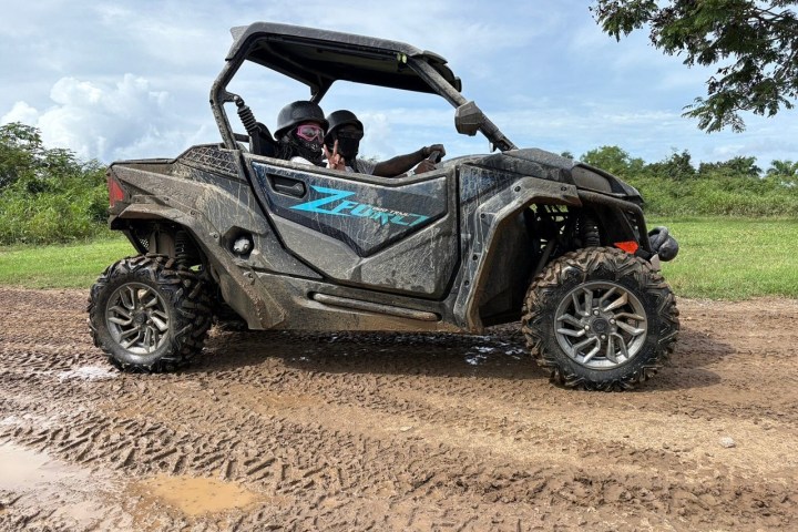 Two people in helmets driving a muddy off-road vehicle on a dirt path.