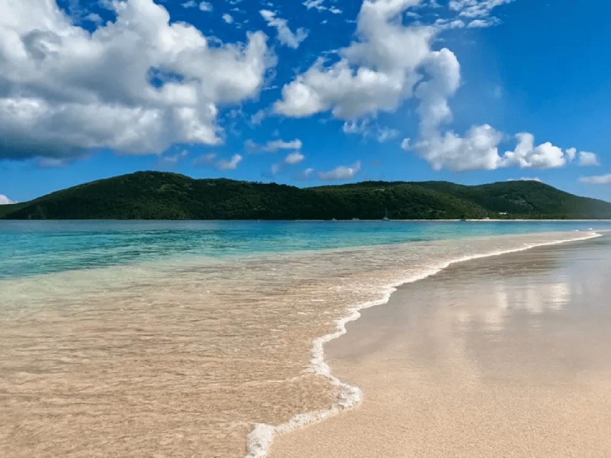 Tropical beach with clear water and green hills under a cloudy blue sky.