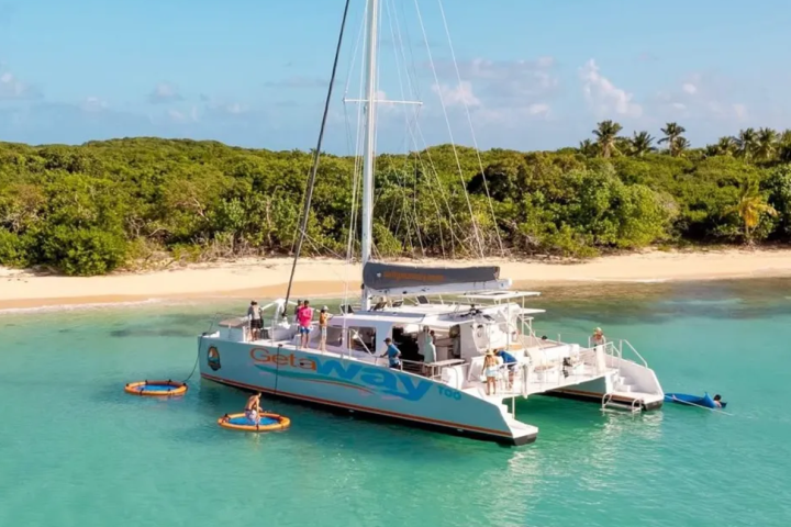 Catamaran anchored near tropical beach with lush greenery, people swimming and using water floats.