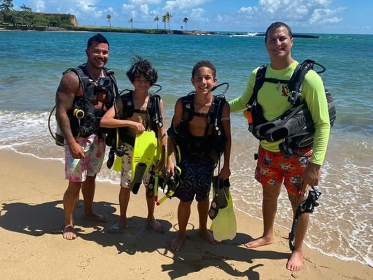 Four people in scuba gear standing on a sandy beach with the ocean in the background.
