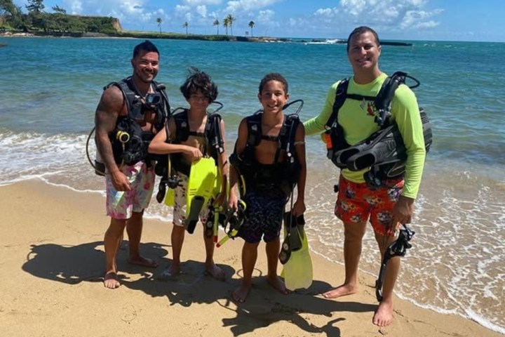 Four people in scuba gear standing on a sandy beach with the ocean in the background.