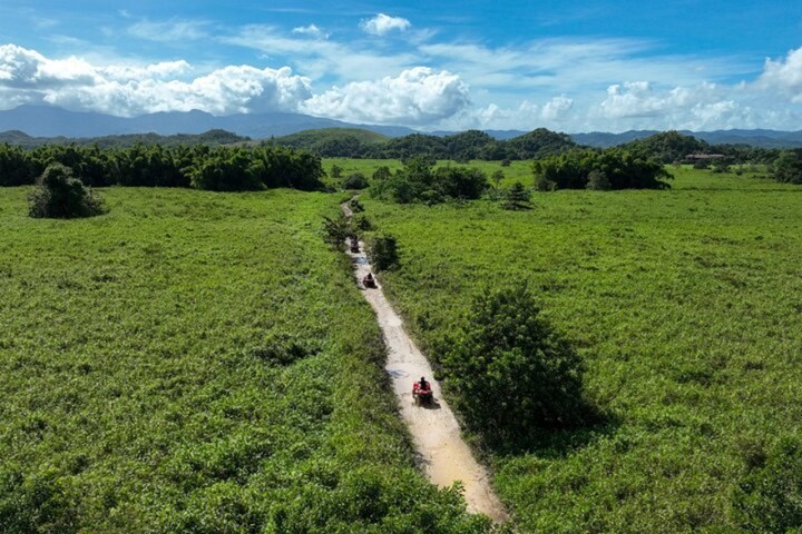 ATVs on a dirt path through green fields with hills and cloudy sky in the background.