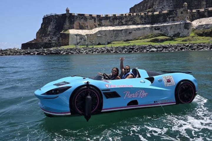 Two people in a blue car-shaped boat on water by a historic fort.