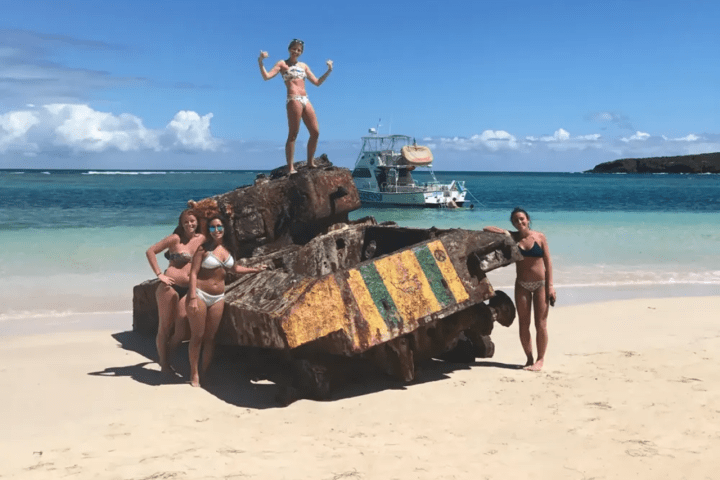 Four people in swimwear posing on and around a rusted tank on a beach with a boat in the background.