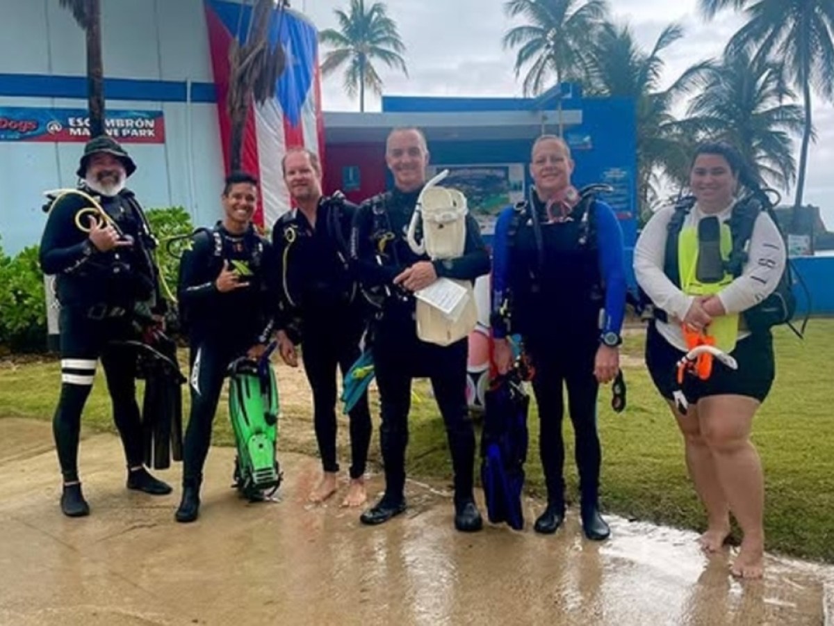 Group of people in scuba gear standing outside near palm trees and a building.